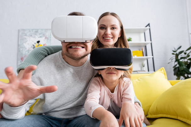 Ребенок и отец играют в гарнитуру VR рядом с улыбающейся мамой дома  - Фото, изображение