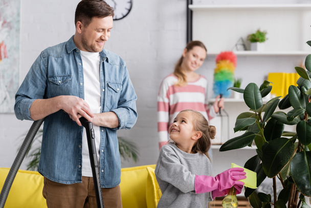 Girl cleaning plant and looking at father with vacuum cleaner at home  - Photo, Image