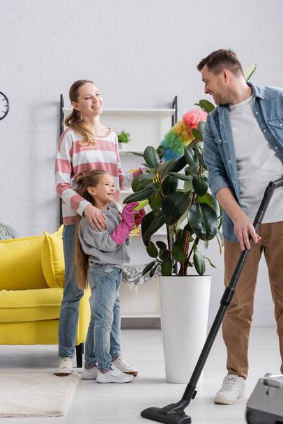 Smiling man with vacuum cleaner looking at family near plant at home  - Photo, Image
