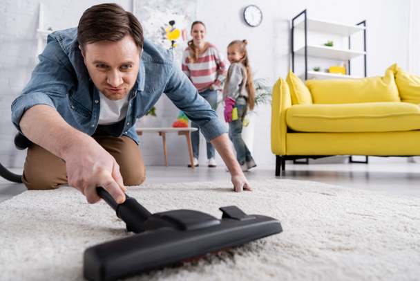 Homme nettoyage tapis sur le premier plan flou près de la famille floue à la maison  - Photo, image