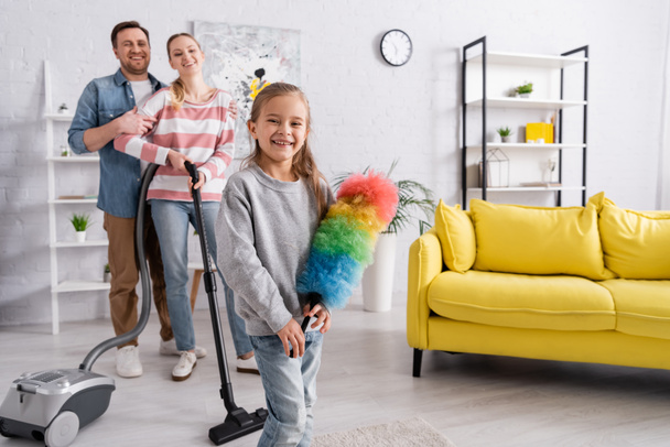 Happy kid holding dust brush near mother with vacuum cleaner and father at home  - Photo, Image