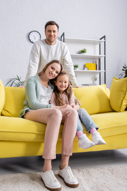 Donna abbracciare il bambino sul divano giallo vicino al marito sorridente  - Foto, immagini