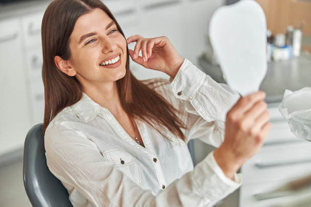 Щаслива молода жінка посміхається перевіряючи свої ідеальні здорові зуби у дзеркалі, сидячи в стоматологічному кріслі в стоматологічному кабінеті
 - Фото, зображення