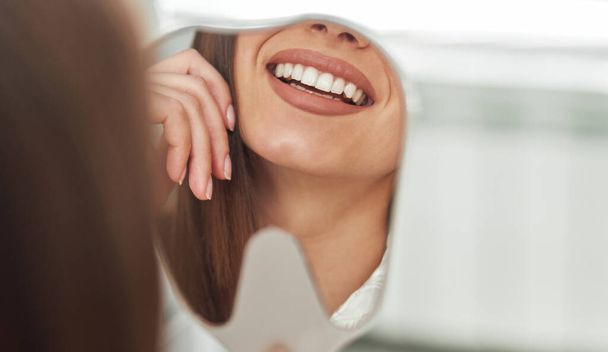 Счастливая молодая женщина улыбается, глядя на свои идеальные здоровые зубы в зеркале крупным планом, в кабинете дантиста - Фото, изображение