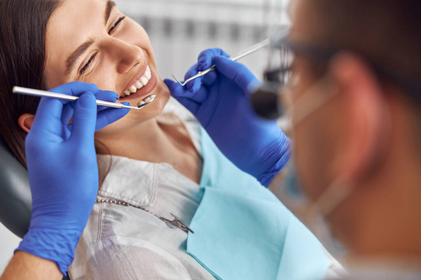Veselý pozitivní zubař a klient v zubařství. Pacientka na zubním zákroku, lékařka na zubních přístrojích v moderní zubní klinice, zblízka. - Fotografie, Obrázek