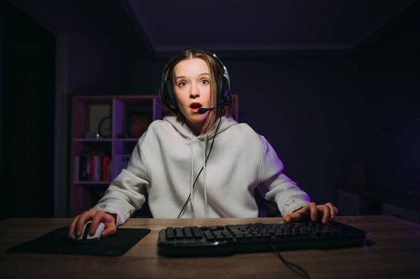 Шокована жінка-гравець грає вдома на комп'ютері в навушниках і дивлячись на камеру зненацька. Емоційна леді поспішає онлайн вдома для гри
. - Фото, зображення