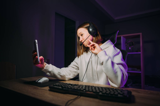 Piękna pani Srimer siedzi w nocy przy komputerze w słuchawkach i komunikuje się z abonentami na aparacie smartfona, grając w gry online. - Zdjęcie, obraz