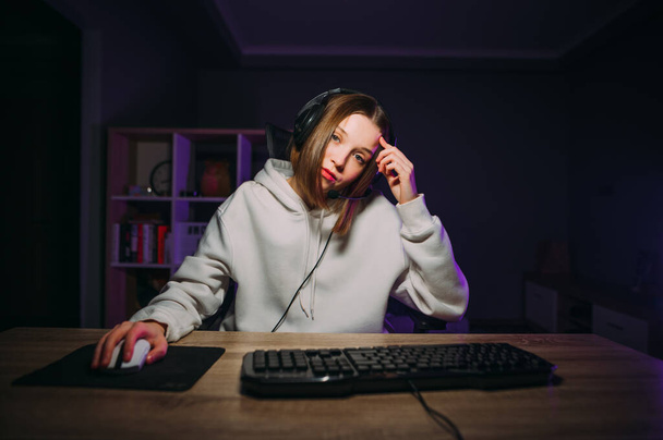 Tired female streamer playing games at night on the computer in the bedroom with a serious face looking at the camera. Lady gamer in white hoodie stream game. - Photo, Image