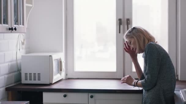 Douloureuse vieille femme solitaire penchée sur le comptoir de la cuisine et regardant par la fenêtre - Séquence, vidéo