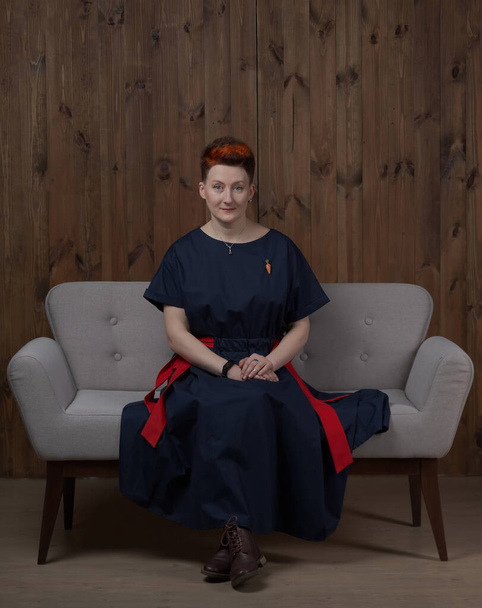Молода жінка блогер або тренер у блакитній сукні позує в студії на сірому дивані
. - Фото, зображення
