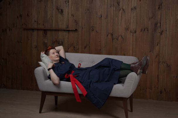 Молодая женщина-блогер или тренер в синем платье позирует в студии на сером диване. - Фото, изображение