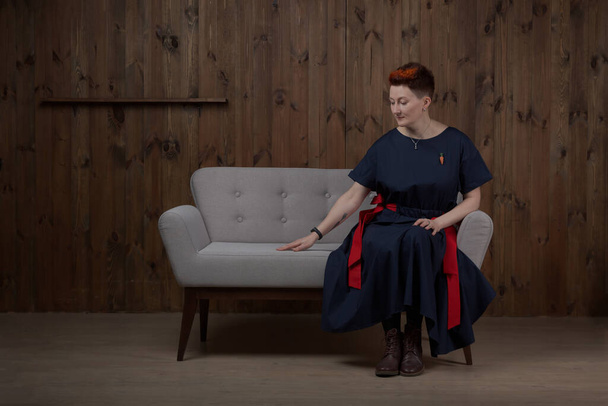 Jovem blogueira ou treinadora em um vestido azul posando no estúdio em um sofá cinza. - Foto, Imagem