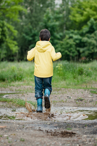 wet child jumping in puddle. Fun on street. Tempering in summer. Splashes, drops of water, outdoor. waterproof boots jump in puddle and mud in rain. Happy childhood - Photo, image