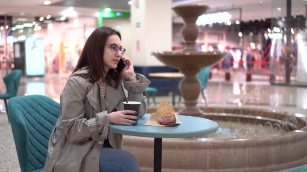Een jonge vrouw in een café spreekt aan de telefoon tegen de achtergrond van een fontein. Decoratie in het café. - Video