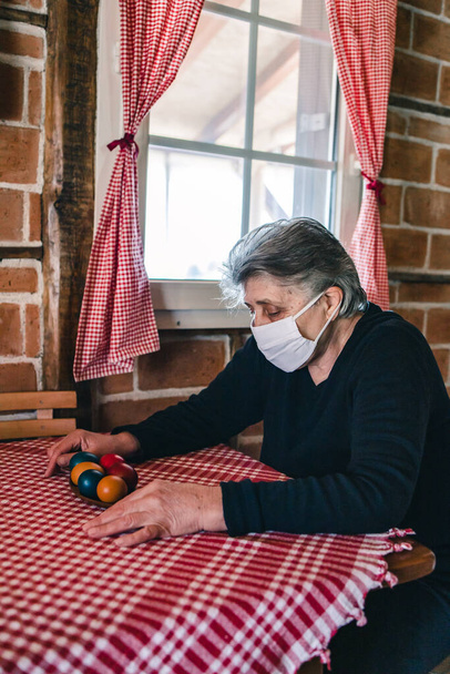 Elderly woman, grandma wearing a mask sitting inside the house by the window alone, looking sad and worried. - Foto, immagini