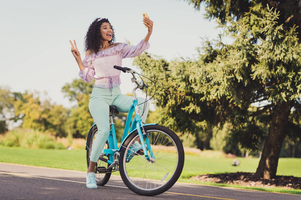 Full length photo of happy afro american brunette woman ride bike in park make selfie v-sign phone outdoors in countryside - Φωτογραφία, εικόνα