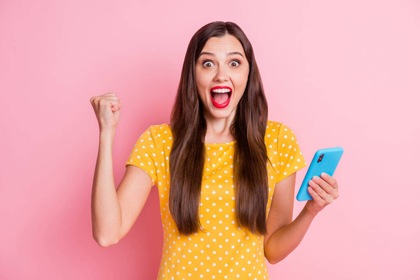 Photo of surprised brown haired lady wear yellow dotted t-shirt raise fist win hold phone isolated on pastel pink color background - Φωτογραφία, εικόνα