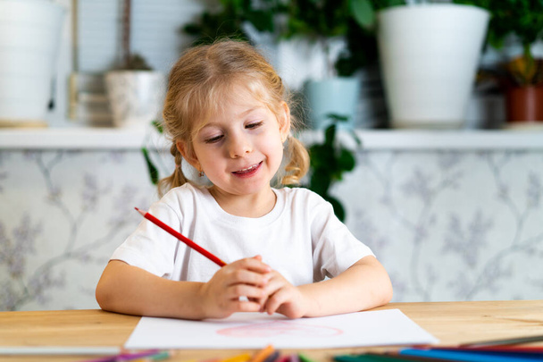 a little blonde girl is sitting at the table, smiling and drawing a heart with a red pencil, there are a lot of colored pencils on the table, learning concept. - Photo, Image