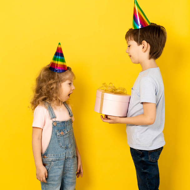 Little blonde girl accepts gift in pink box for her birthday from her brother, on a yellow background, children in caps.  - Fotografie, Obrázek