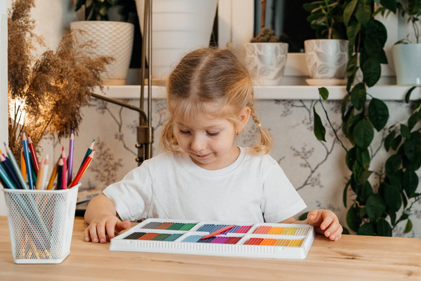 a little blonde girl with a bright colorful palette of colored plasticine for modeling at the table at home, hobbies and child development - Zdjęcie, obraz