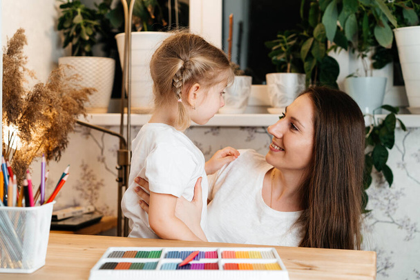 moeder en dochter zitten aan een tafel met gekleurde plasticine voor modellenwerk, kijken elkaar aan en glimlachen.Het concept van Moederdag en Kinderdag - Foto, afbeelding