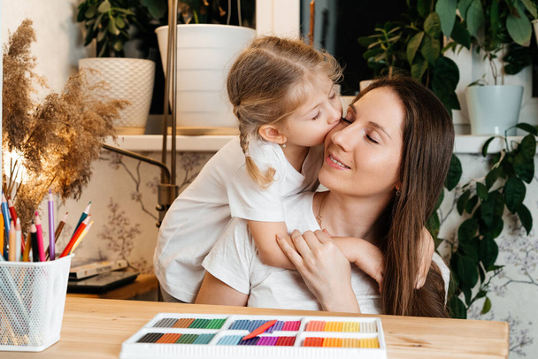 Kızı annesine sarılıyor ve onu öpüyor. Renkli plastiklerin modellik için olduğu masaya oturuyorlar. Anneler Günü ve Çocuklar Günü konsepti - Fotoğraf, Görsel