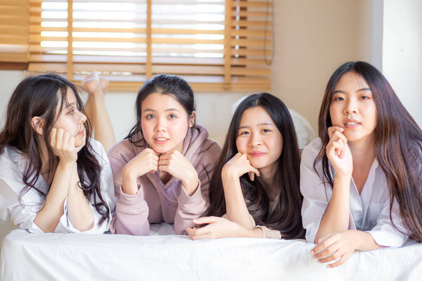 Dört Asyalı kadın yatakta yatıp gülümsüyor. Kameraya bakıyorlar. Mutlu arkadaşlar ya da genç kızlar evdeki rahat odada eğleniyorlar. - Fotoğraf, Görsel