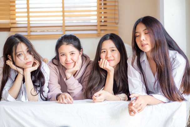 Dört Asyalı kadın yatakta yatıp gülümsüyor. Kameraya bakıyorlar. Mutlu arkadaşlar ya da genç kızlar evdeki rahat odada eğleniyorlar. - Fotoğraf, Görsel