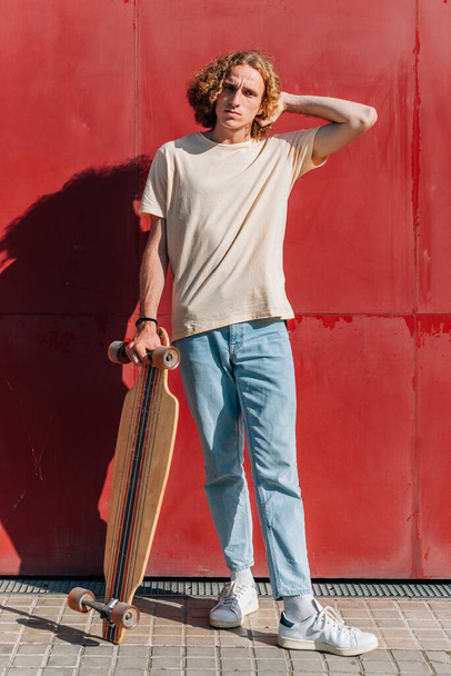 Schöner junger Mann mit langen roten Haaren, der mit seinem Skateboard auf der Straße steht - Foto, Bild