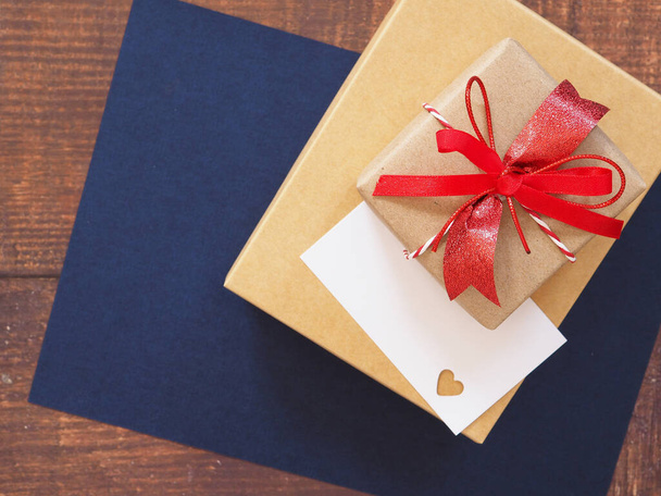 Vista superior, pajarita roja, caja de regalo marrón y tarjeta blanca perforante en forma de corazón sobre papel azul sobre fondo de madera con espacio para copiar - Foto, imagen