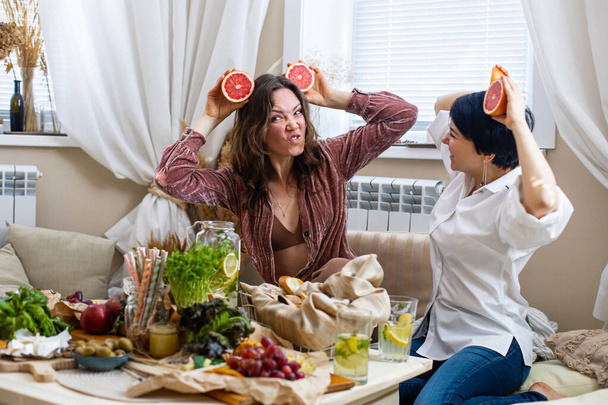 Happy female friends showing tongues posing with halves of red grapefruits on eyes laughing at hen-party. Cheerful woman playing covering face by citrus vitamin having fun. Shot with RED camera in 4K - Φωτογραφία, εικόνα