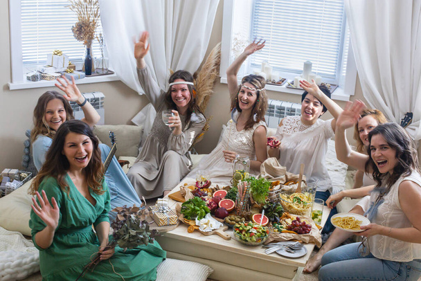 Happy bohemian hippie woman smiling enjoying hen-party at comfortable boho scandi living room. Cheerful female eating healthy food playing guitar gossiping communicating together relaxing at home - Φωτογραφία, εικόνα