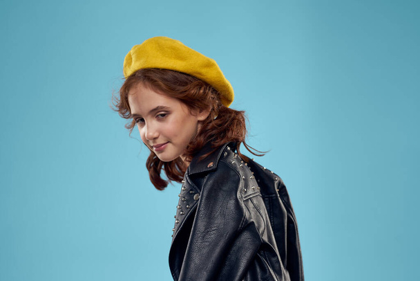 Mavi arka planda siyah deri ceketli ve kafasında sarı şapkalı şık bir kız. - Fotoğraf, Görsel