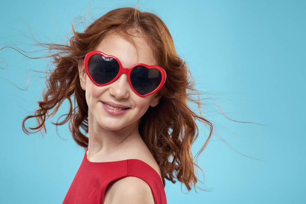 Niña en gafas de sol en forma de corazones vestido rojo pelo rizado fondo azul - Foto, Imagen