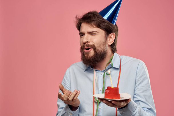 Homme avec un gâteau avec une bougie et une casquette d'anniversaire sur un fond rose - Photo, image