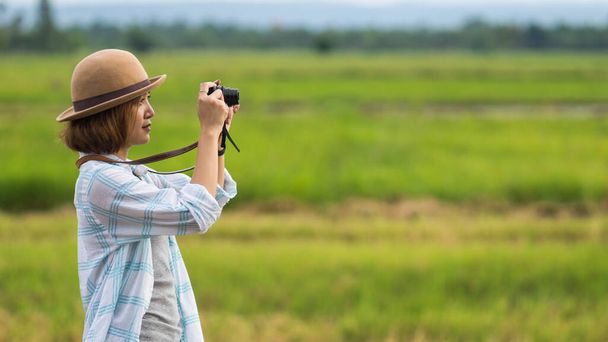 Μια τουρίστρια απολαμβάνει να φωτογραφίζει το τοπίο κατά τη διάρκεια των καλοκαιρινών διακοπών της στον τομέα του πράσινου. Μια κομψή ταξιδιώτισσα γυναίκα με καπέλο που φωτογραφίζει τον εαυτό της και το τοπίο ενώ χαλαρώνει - Φωτογραφία, εικόνα