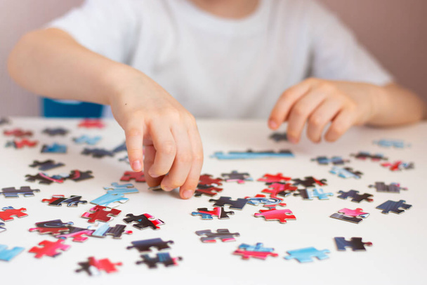 The child collects a puzzle. Elements of the puzzle are laid out on a white table. Children's hands during the game. Home leisure. - Photo, Image