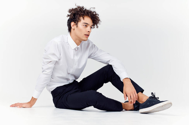 curly-haired guy in shirt and trousers and sneakers sits on the floor indoors side view - Photo, Image