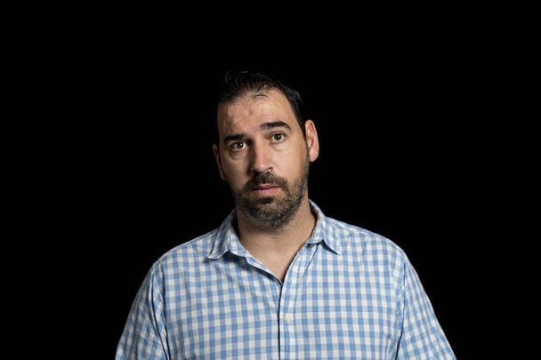 Портрет бородатого мужчины, одетого в белую и синюю клетчатую рубашку, создающий беспокойство изолированного на черном фоне студии. Концепция реального человека - Фото, изображение