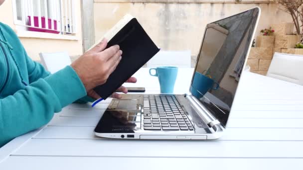 Junger Mann arbeitet mit seinem Laptop im Garten seines Hauses und schreibt in ein Notizbuch - Filmmaterial, Video