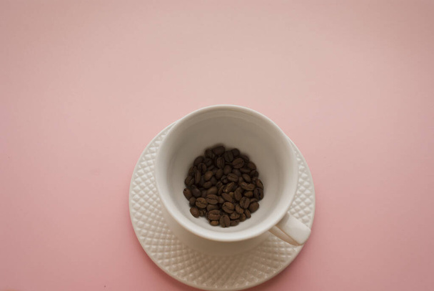 Taza blanca y platillo con granos de café negro sobre un fondo rosa brillante - Foto, imagen