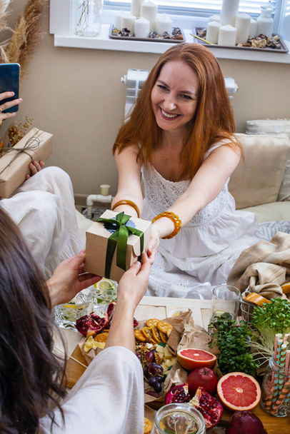 Adorable rejoicing redhead woman taking wrapped gift box celebrating birthday in modern hippie bohemian style. Happy female receiving present from friends having positive emotion at hen-party - Foto, Bild