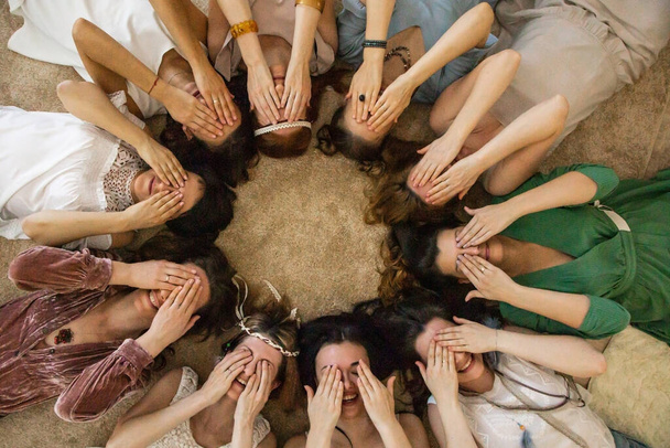 Group of funny fashion female friends posing covering eyes by hands lying circle enjoying friendship. Cheerful stylish hippie woman having fun at hen-party spending time together hiding by palms - 写真・画像