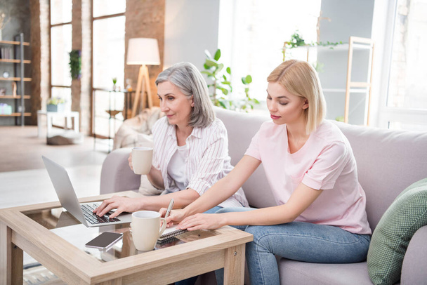 Photo of serious family grey haired mom and blonde daughter sit sofa write note use laptop indoors inside house home - Foto, Bild