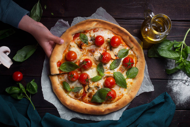 Pizza with tomatoes, mushroom and basil on a dark background. Boy takes a piece of pizza - Zdjęcie, obraz