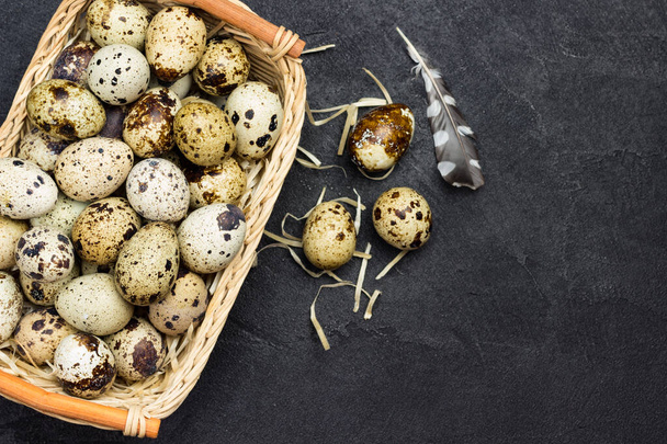 Quail eggs in wicker basket. Egg and feather on table. Black background. Flat lay. Copy space - Fotografie, Obrázek