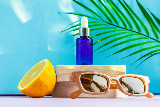 A cosmetic jar with an eyedropper stands on two granite slabs, like on a catwalk, next to half a yellow lemon, a palm leaf, sunglasses and the glare of the sun in the background. Cosmetics for summer. - Foto, Imagen