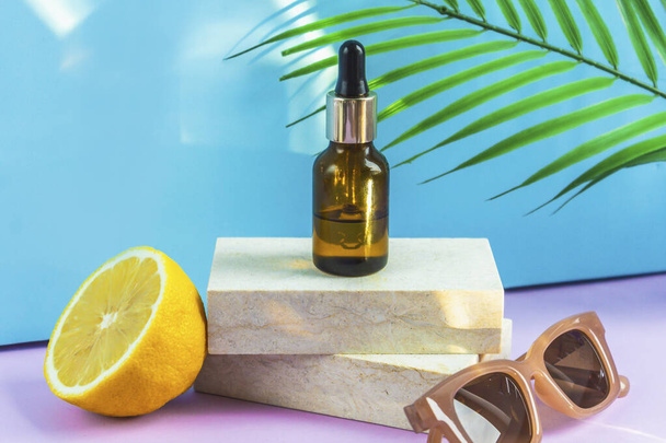 A cosmetic jar with an eyedropper stands on two granite slabs, like on a catwalk, next to half a yellow lemon, a palm leaf, sunglasses and the glare of the sun in the background. Cosmetics for summer. - Foto, immagini