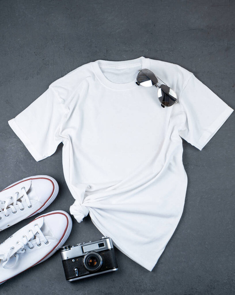 White T-shirt with place for text. Sneakers, glasses, camera - a set of clothes for walking. T-shirt mockup for printing. Stylish kit. Grey background with copy space. Side knot. - Foto, Bild