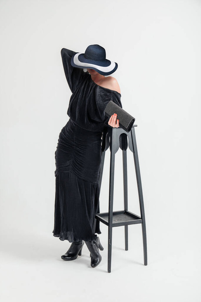 Hermosa joven con una hermosa figura en un vestido oscuro con sombrero posando sobre un fondo blanco en el estudio. - Foto, Imagen